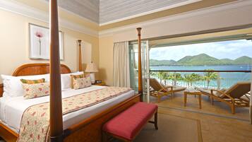 A hotel room with a bed, a bench, a table, and a balcony with a view of the sea and hills.
