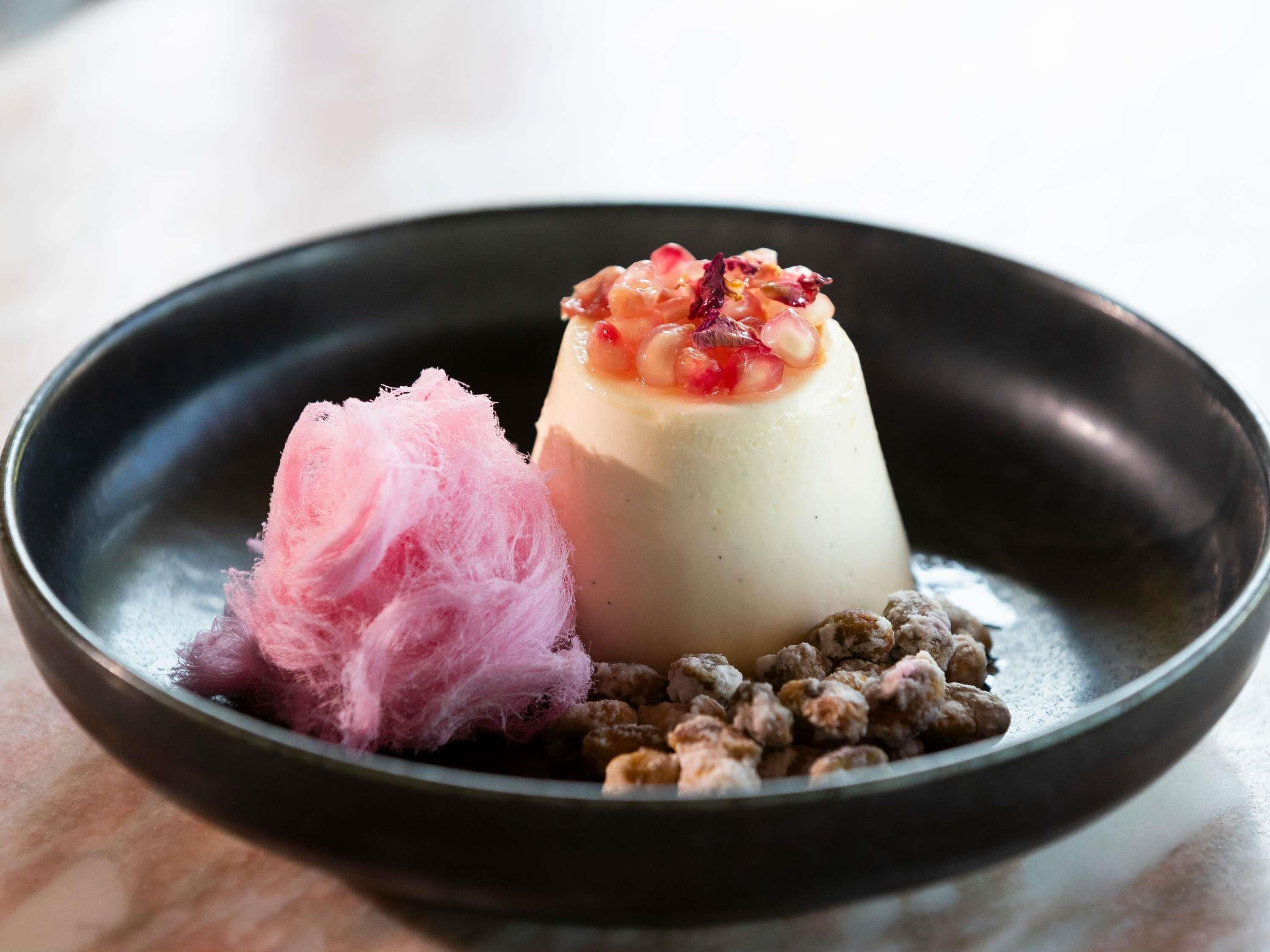 Un postre con base blanca cubierto de fruta roja y un elemento esponjoso rosado sobre un plato oscuro.
