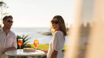 A couple seated at an outdoor table with drinks and a plate of food, overlooking a body of water.