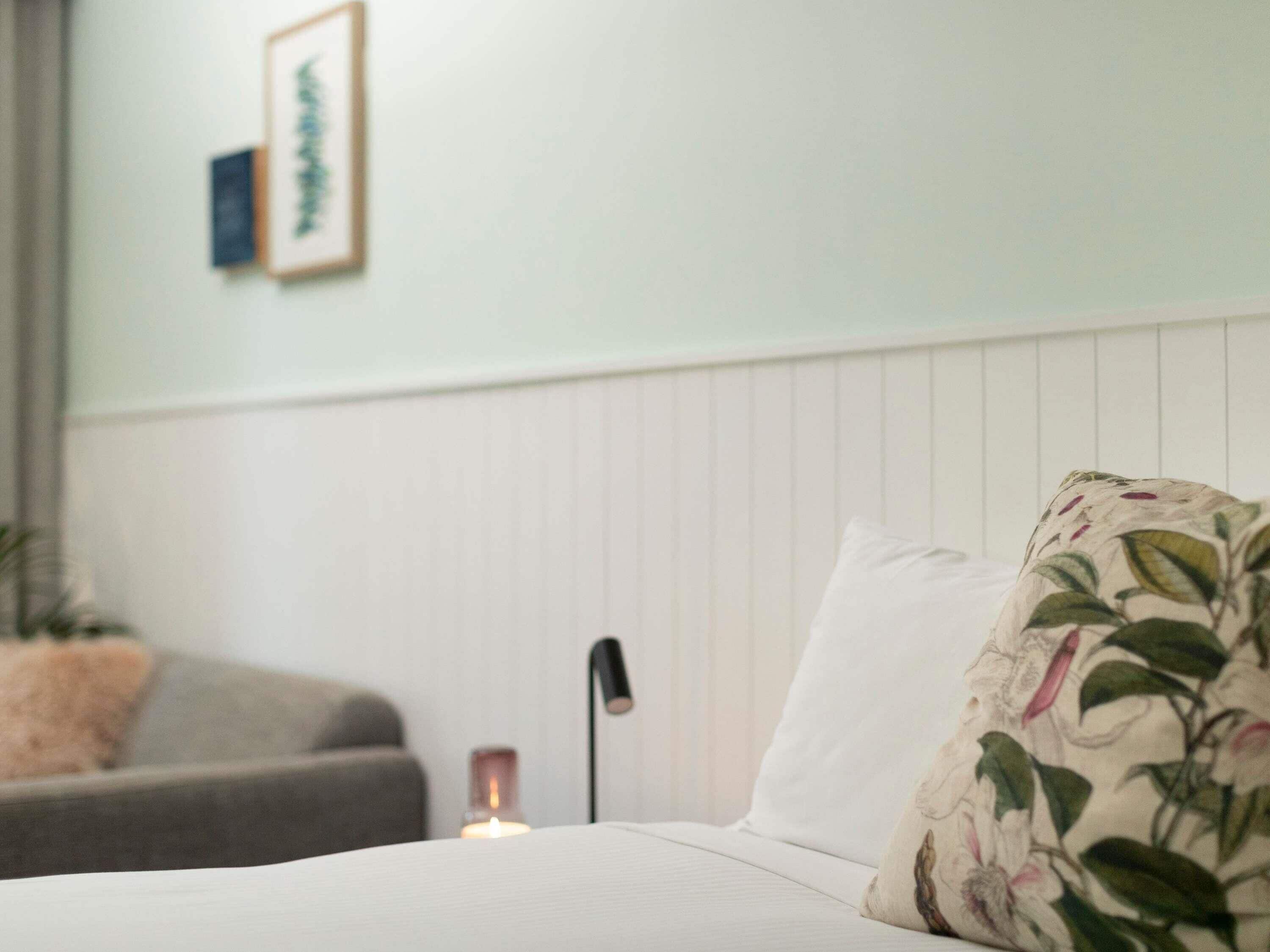 A bedroom with a bed, a bedside table with a candle, a wall-mounted picture, and a grey sofa in the background.