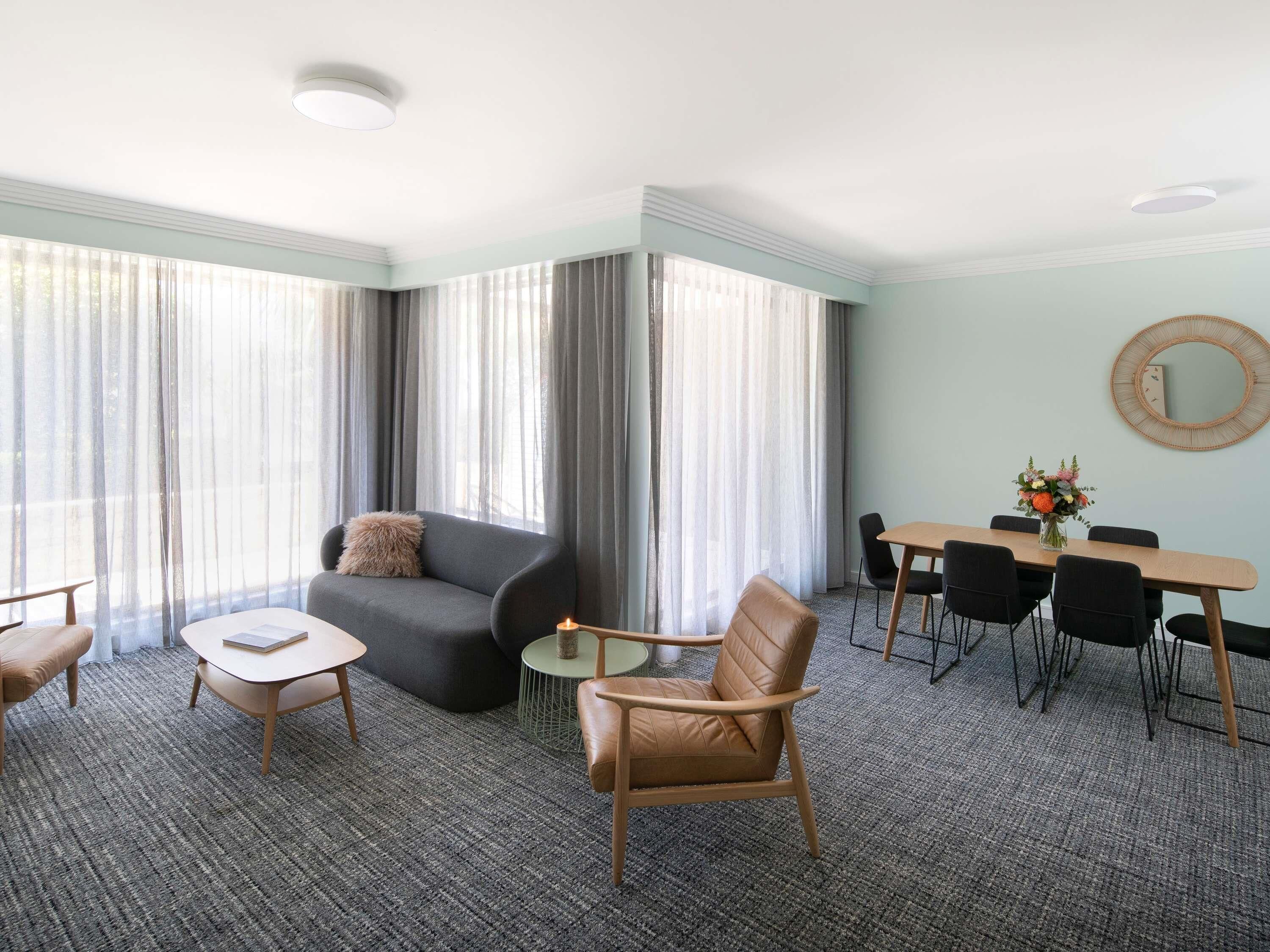 Een hotelkamer met een bank, een klein rond tafeltje, een eettafel met stoelen en een grote spiegel aan de muur.