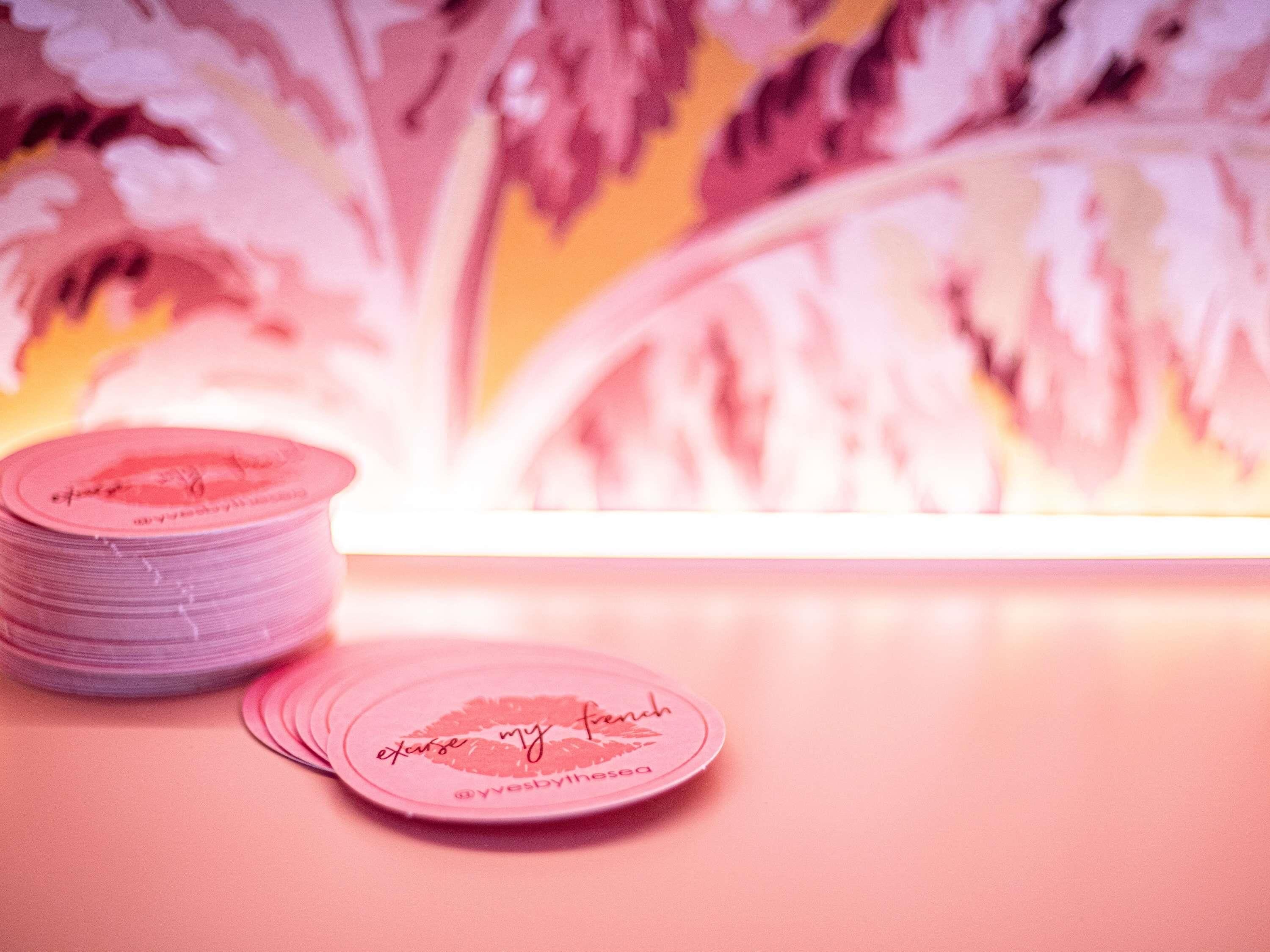 A stack of pink coasters with a logo and text, placed on a table with a floral patterned background.