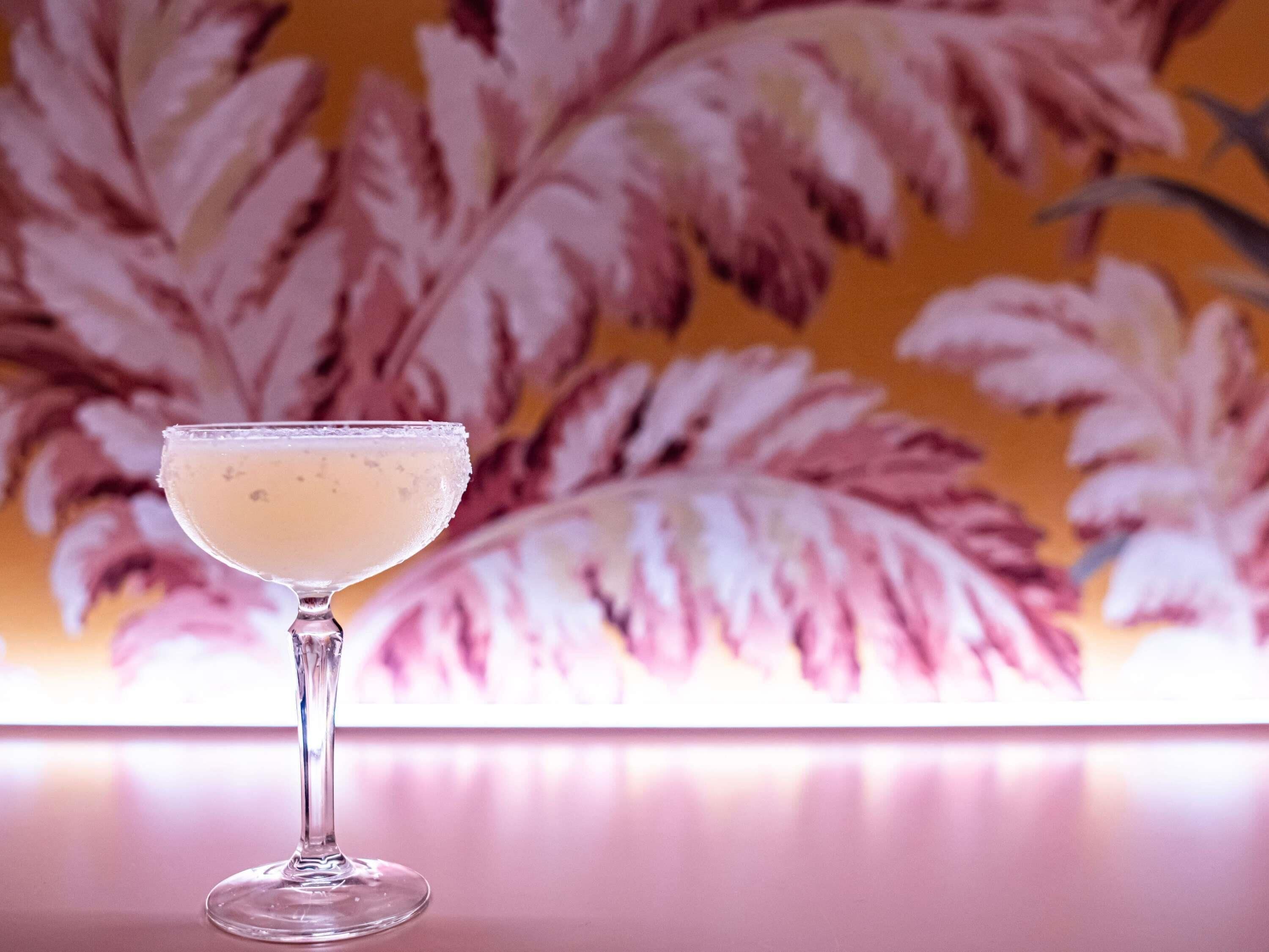 En cocktail i et stilkskank med en reflekterende overflade og blomstret tapet i baggrunden.
