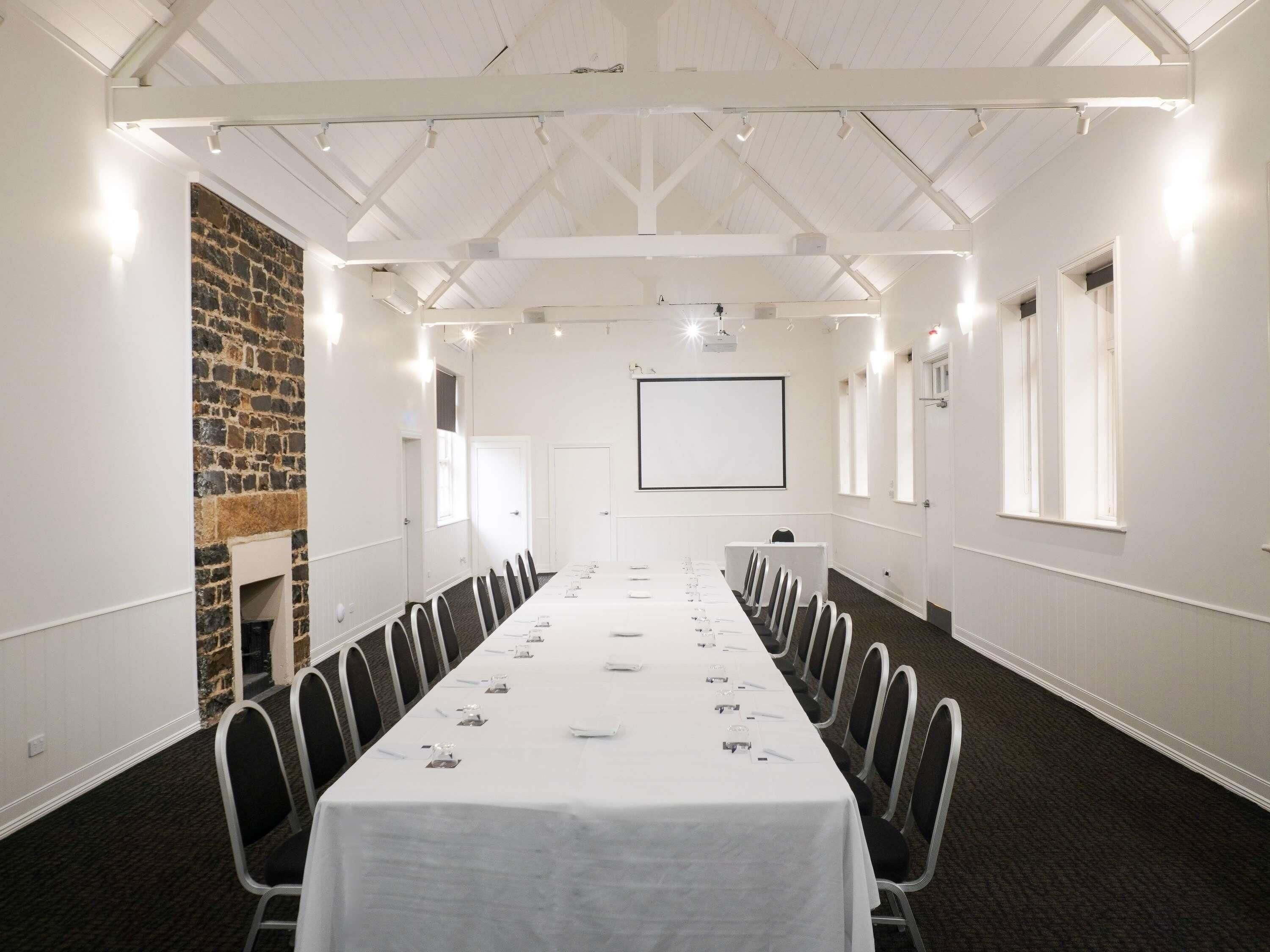 A conference room with a long table, chairs, a projector screen, and a stone wall.