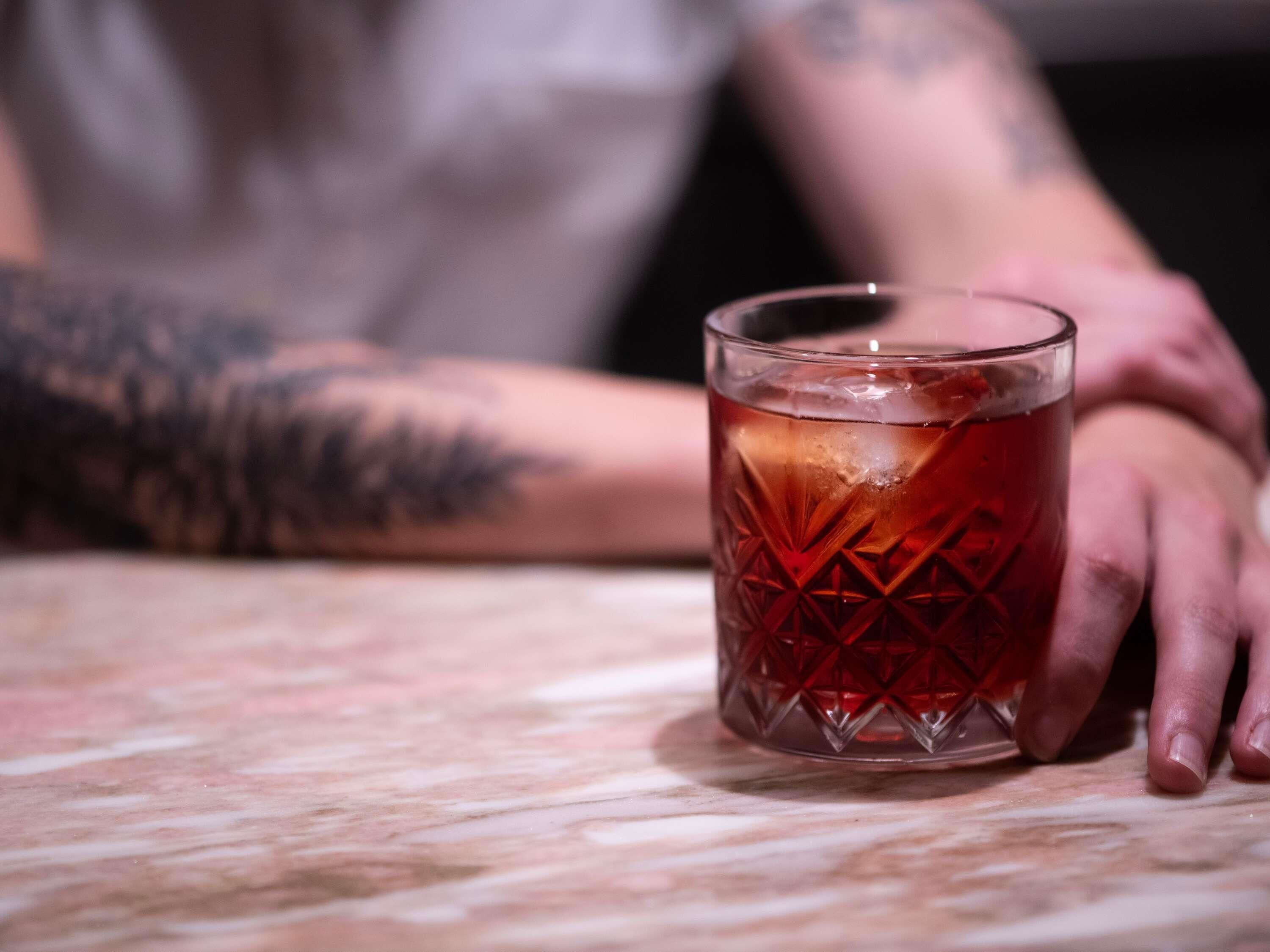 A glass of amber liquid on a marble surface with a person's arm resting beside it.