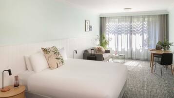 A hotel room with a large bed, a desk, a chair, a small table, a lamp, and a potted plant.