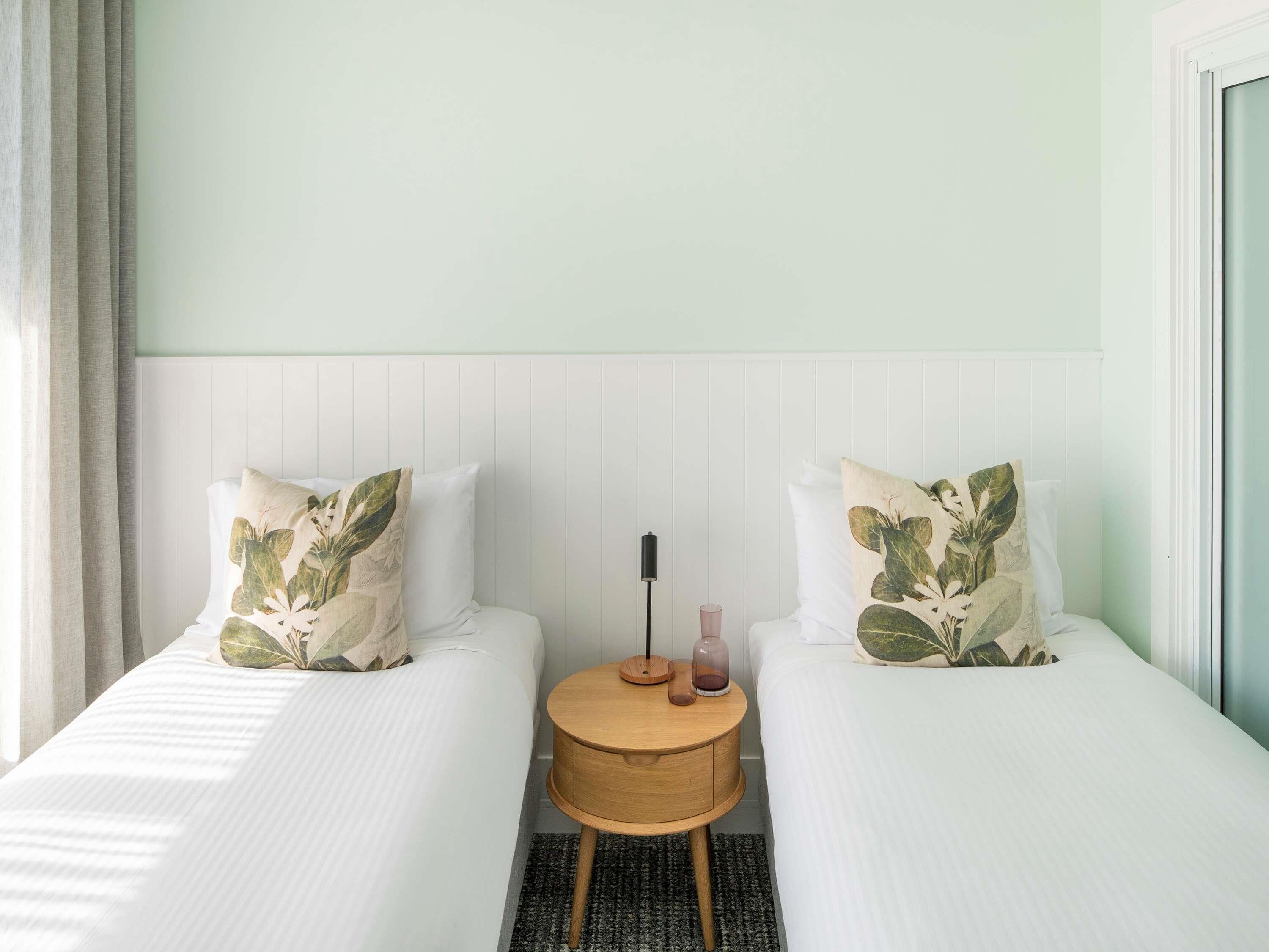 Une chambre d’hôtel avec deux lits simples, une table de chevet ronde en bois et une tête de lit blanche.