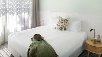 A hotel room with a bed, a bedside table with a lamp, a hat on the bed, and a window with curtains.