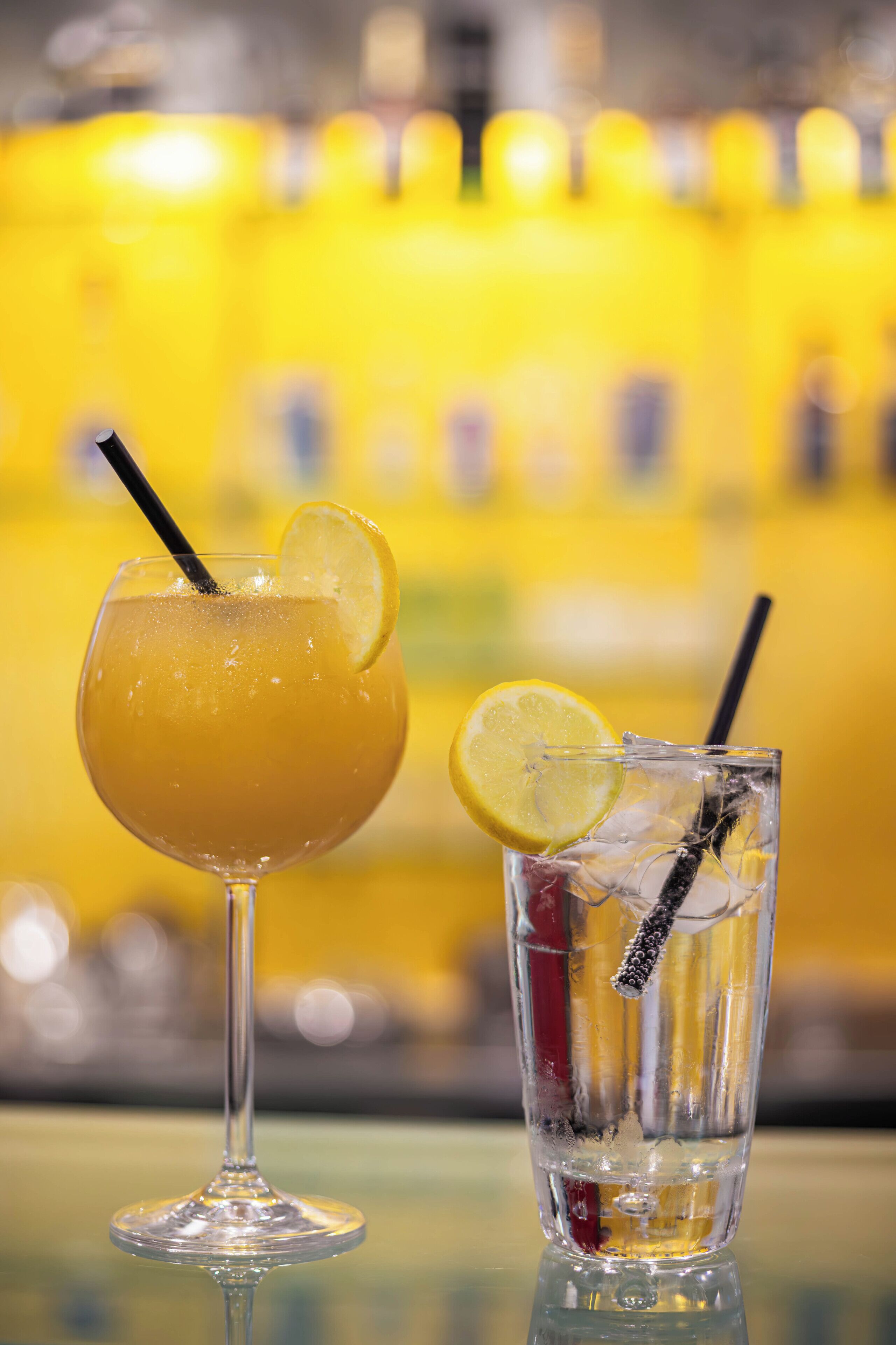Two drinks, one in a stemmed glass and one in a tall glass, with straws and lemon slices.