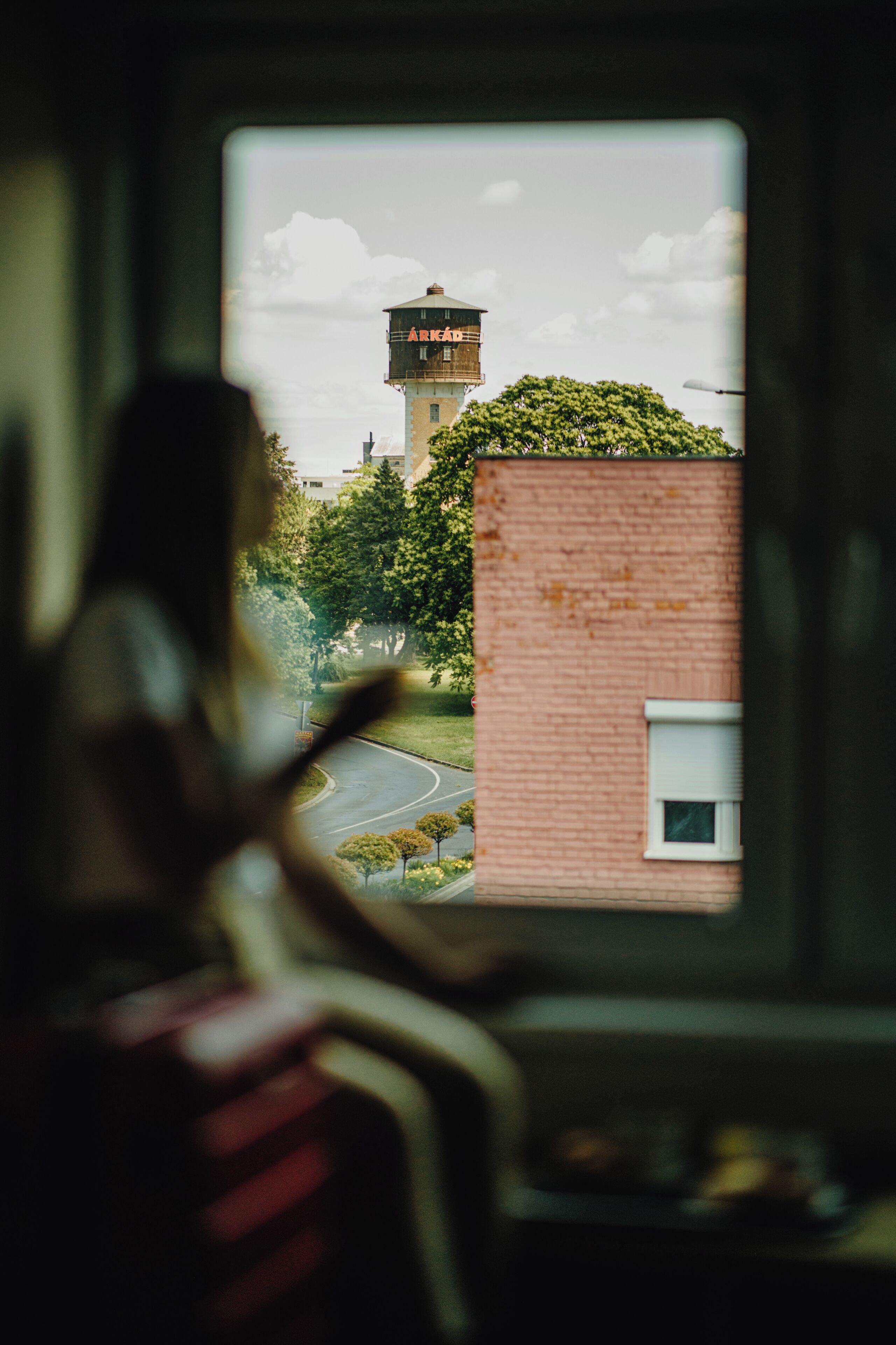 Une personne regarde par une fenêtre un château d’eau et un bâtiment en briques.