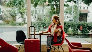 Uma pessoa sentada à mesa com uma mala, uma cadeira e uma janela com vista para edifícios e árvores.