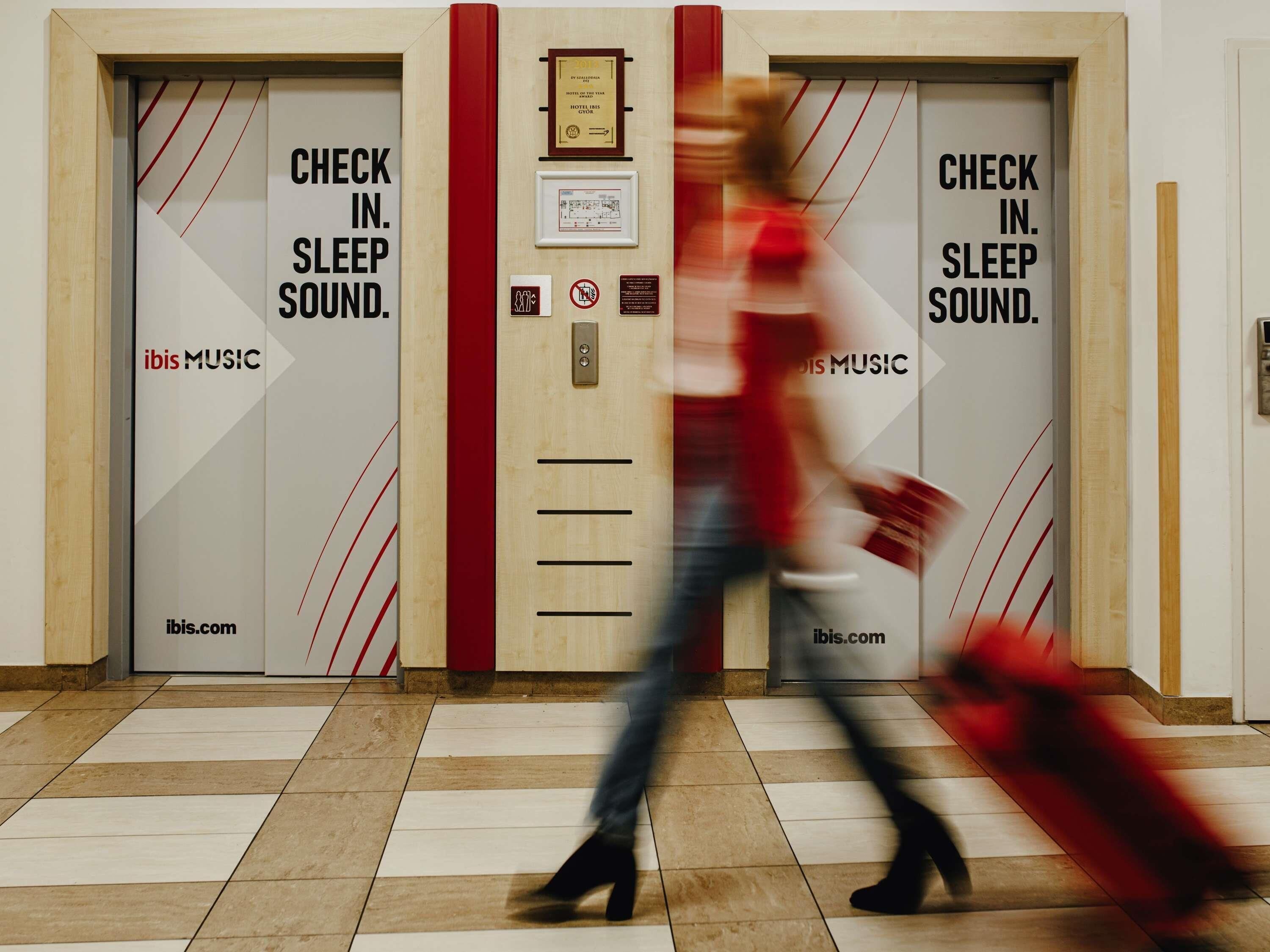 Deux portes d’ascenseur avec les inscriptions « ENREGISTREMENT. PASSEZ UNE BONNE NUIT » et « ibis MUSIC », une plaque et une bande verticale rouge.