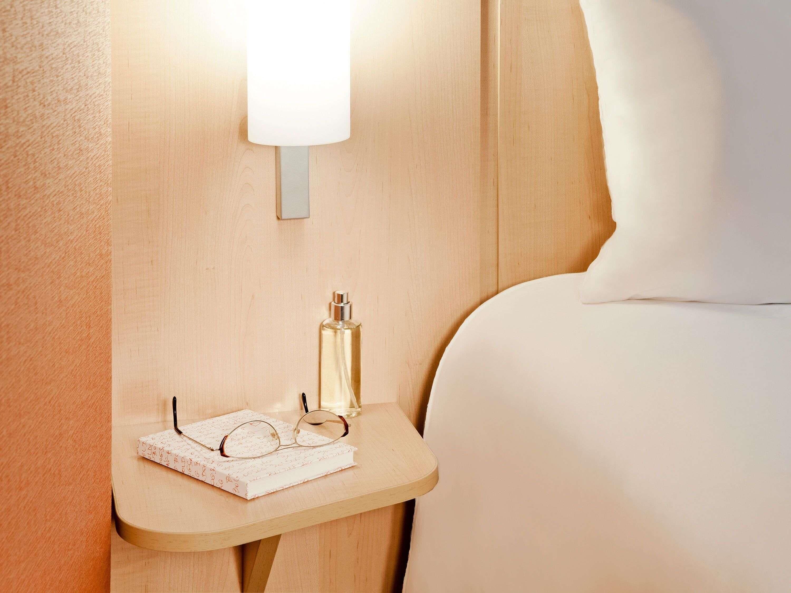 Un lit bien fait, avec un oreiller blanc, une table de chevet sur laquelle se trouvent une lampe, une bouteille, des lunettes et un carnet.