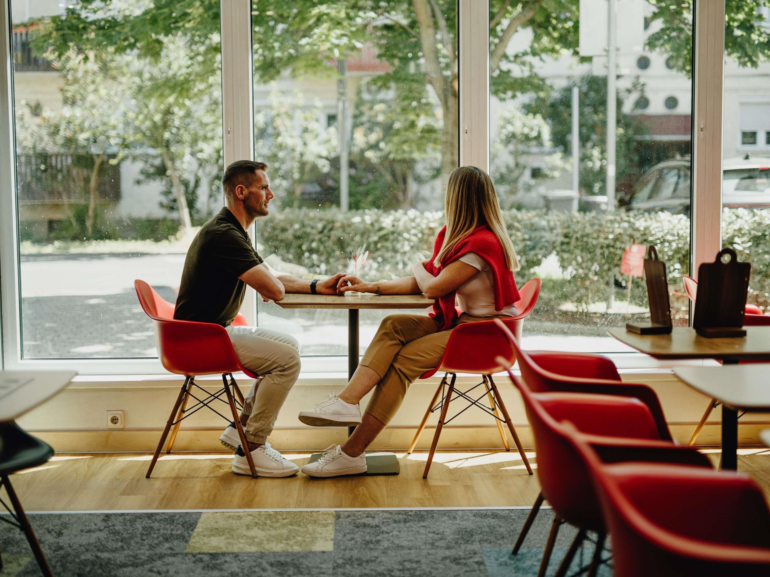 A couple seated at a table inside a cafe with large windows, wooden flooring, and red chairs.