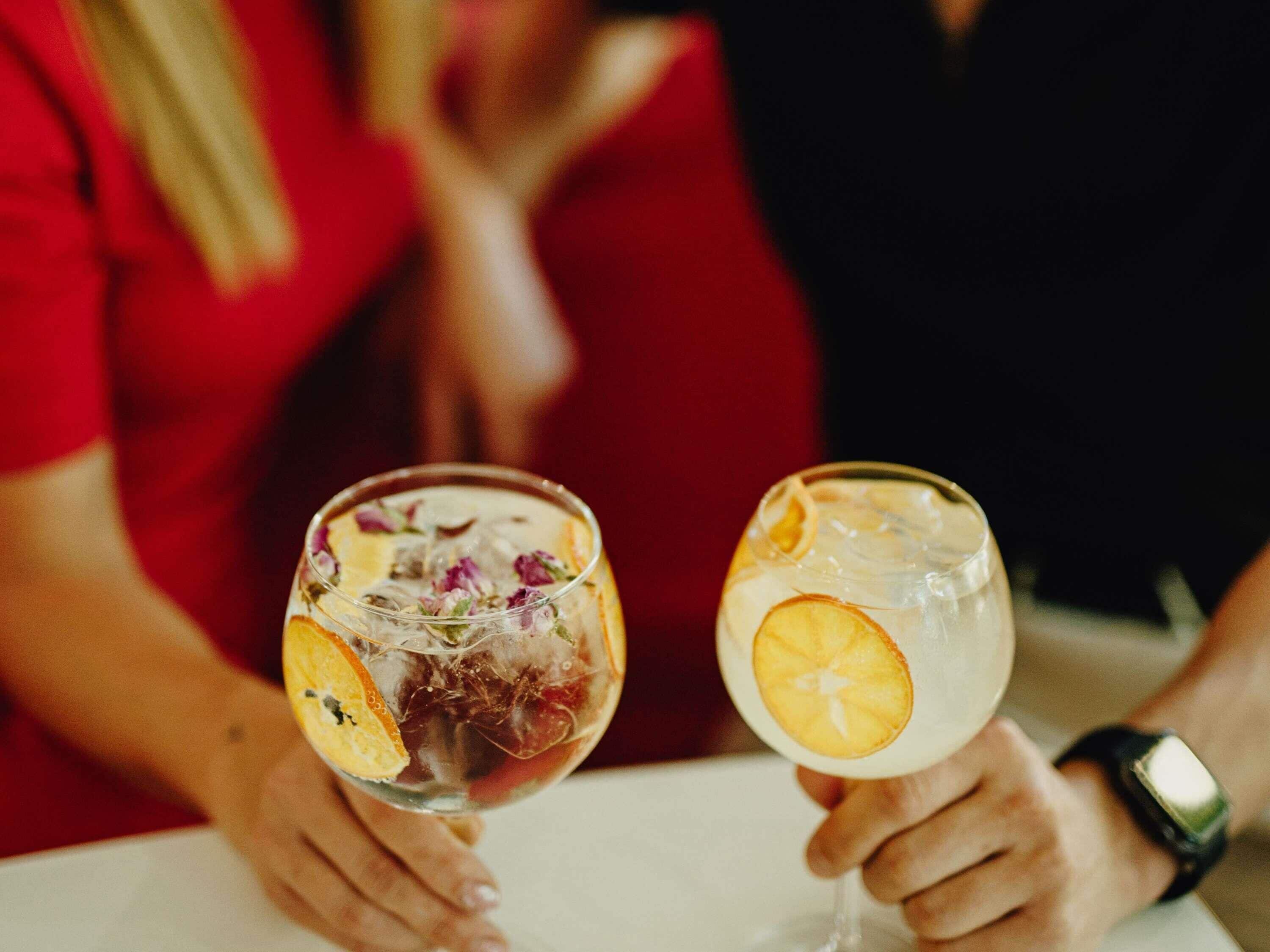 Twee personen houden glazen met een helder drankje, ijs en citruspartjes.