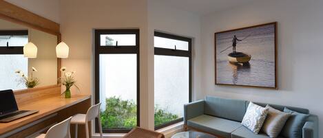 Una sala de estar moderna con un escritorio de madera, sillas blancas, una mesa de centro de vidrio, un sofá azul y un ventanal con vistas a la vegetación.