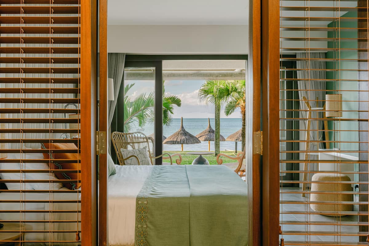 Une chambre avec un lit, une chaise et une vue sur la plage et l’océan, visible par une fenêtre munie de stores.