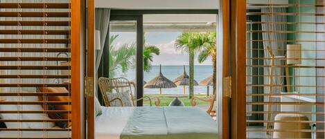 A room with a bed, a chair, and a view of the beach and ocean through a window with blinds.