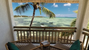 A balcony with wicker furniture, a small table, and a view of a beach with boats and palm trees. - Veranda Paul & Virginie Hotel & Spa - Adults Only (Grand Gaube)