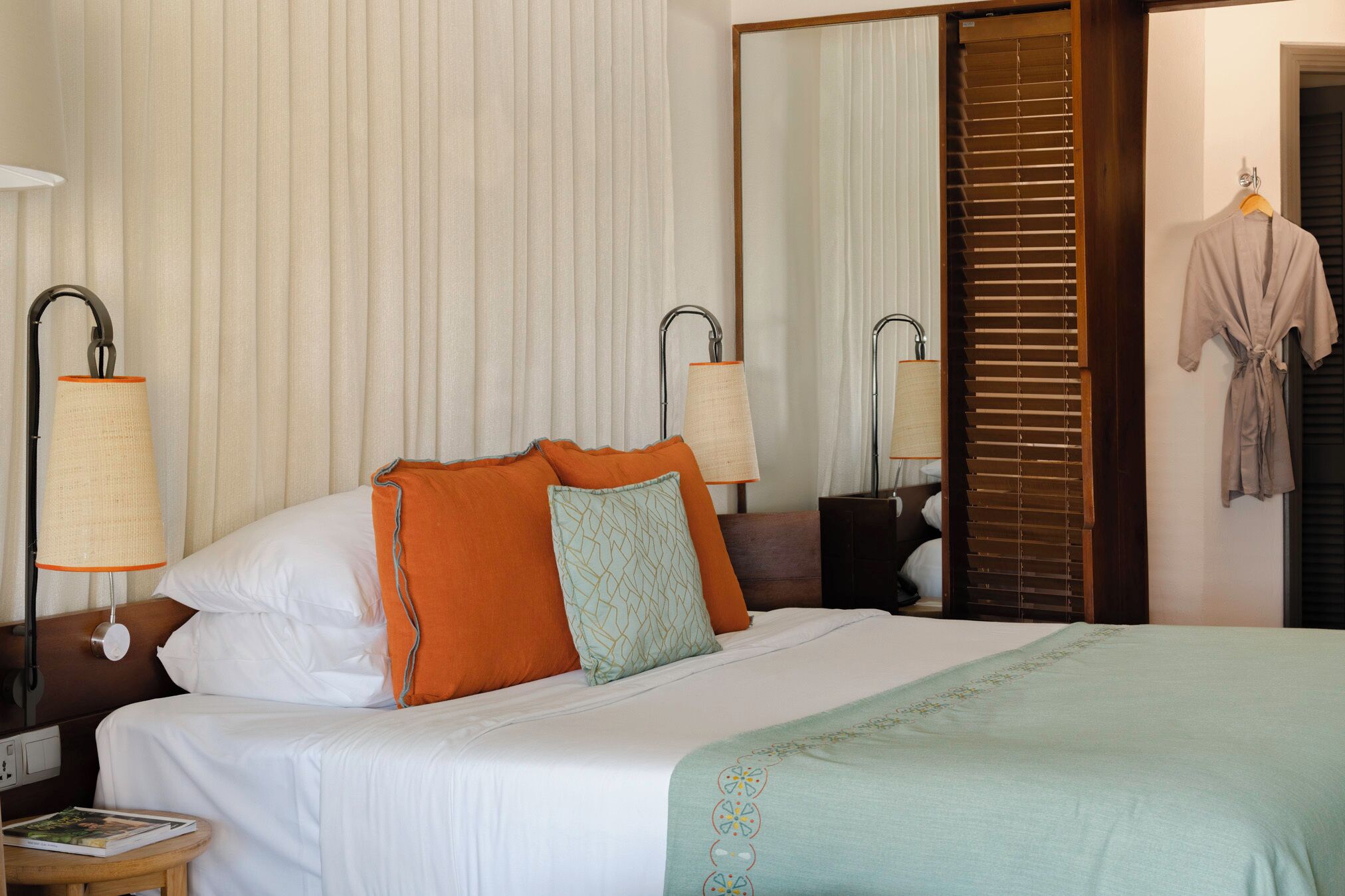 A hotel room with a neatly made bed, two bedside lamps, a large mirror, and a robe hanging on the door.