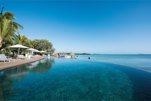 A clear blue pool with lounge chairs and umbrellas, surrounded by palm trees and a wooden deck.