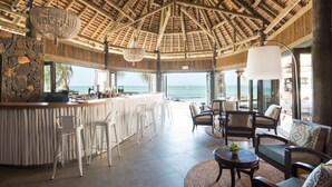 A bar area with a stone wall, white bar stools, and a view of the ocean. There are wicker chairs and a round table with a plant. - Veranda Paul & Virginie Hotel & Spa - Adults Only (Grand Gaube)