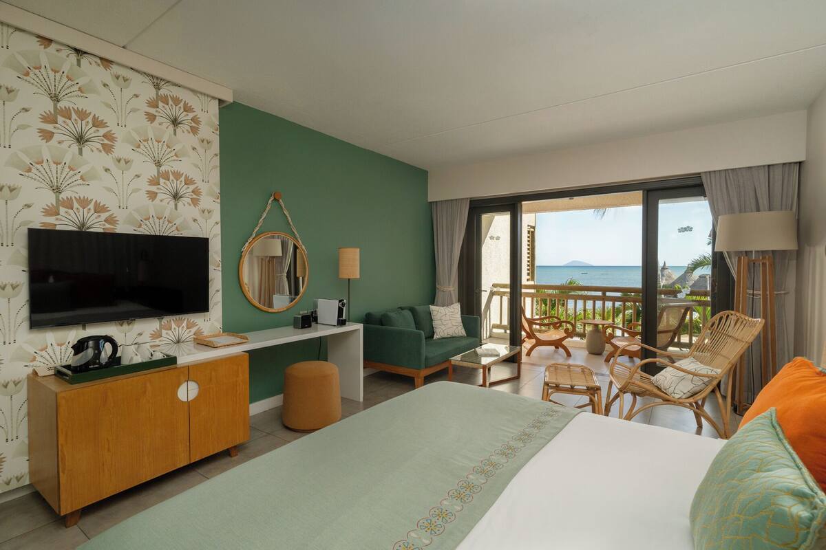 Une chambre d’hôtel comprenant un lit, un bureau, une télévision, un balcon avec coin salon extérieur et une vue sur la mer.