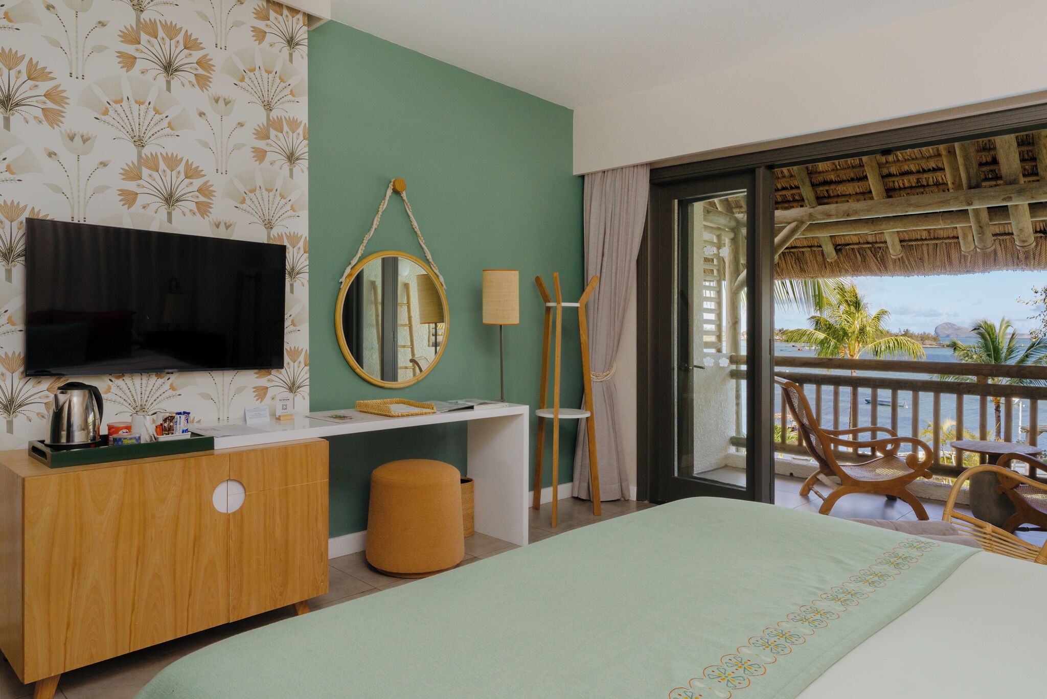 A hotel room with a bed, a TV, a desk, a mirror, a window with a view of the sea, and a balcony with chairs.