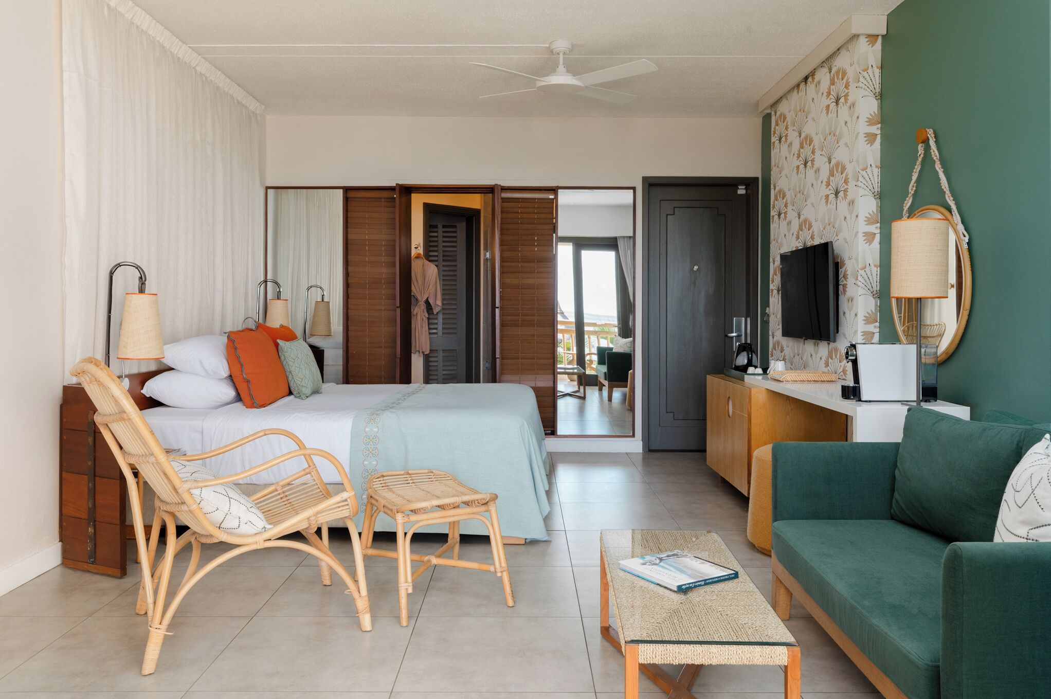 Una habitación de hotel con una cama, dos sillones de mimbre, un sofá verde, una mesita, un televisor y un espejo.