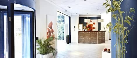 Et hotellobby med receptionsdisk, potteplanter og et stort vindue.