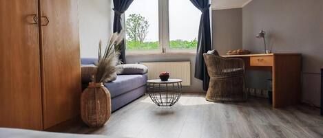 Una habitación con cama, sofá, escritorio, silla y ventana con cortinas, y vista a zonas verdes.
