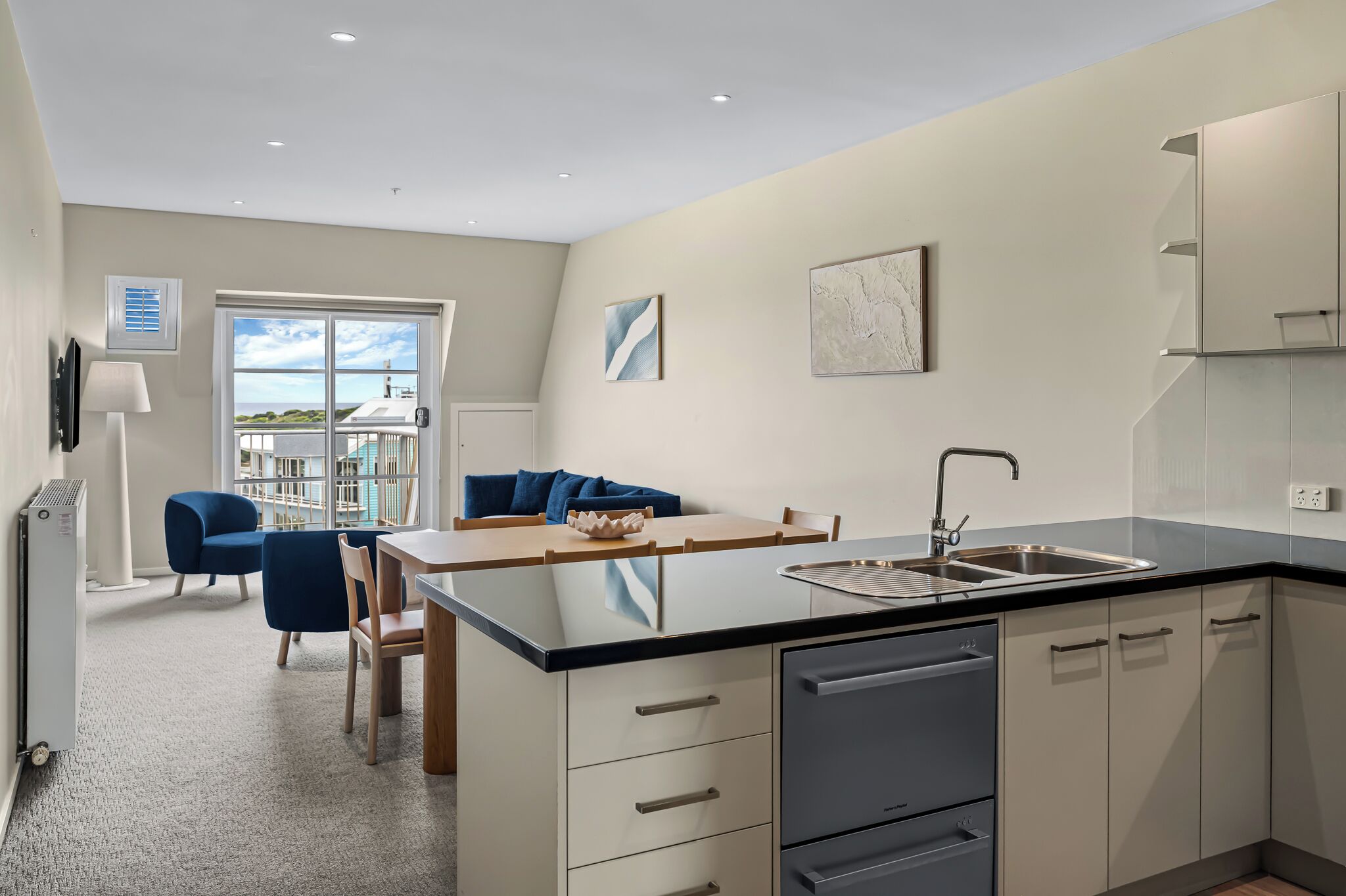 A modern kitchen with a central island, a dining area with blue chairs, and a view through a large window.