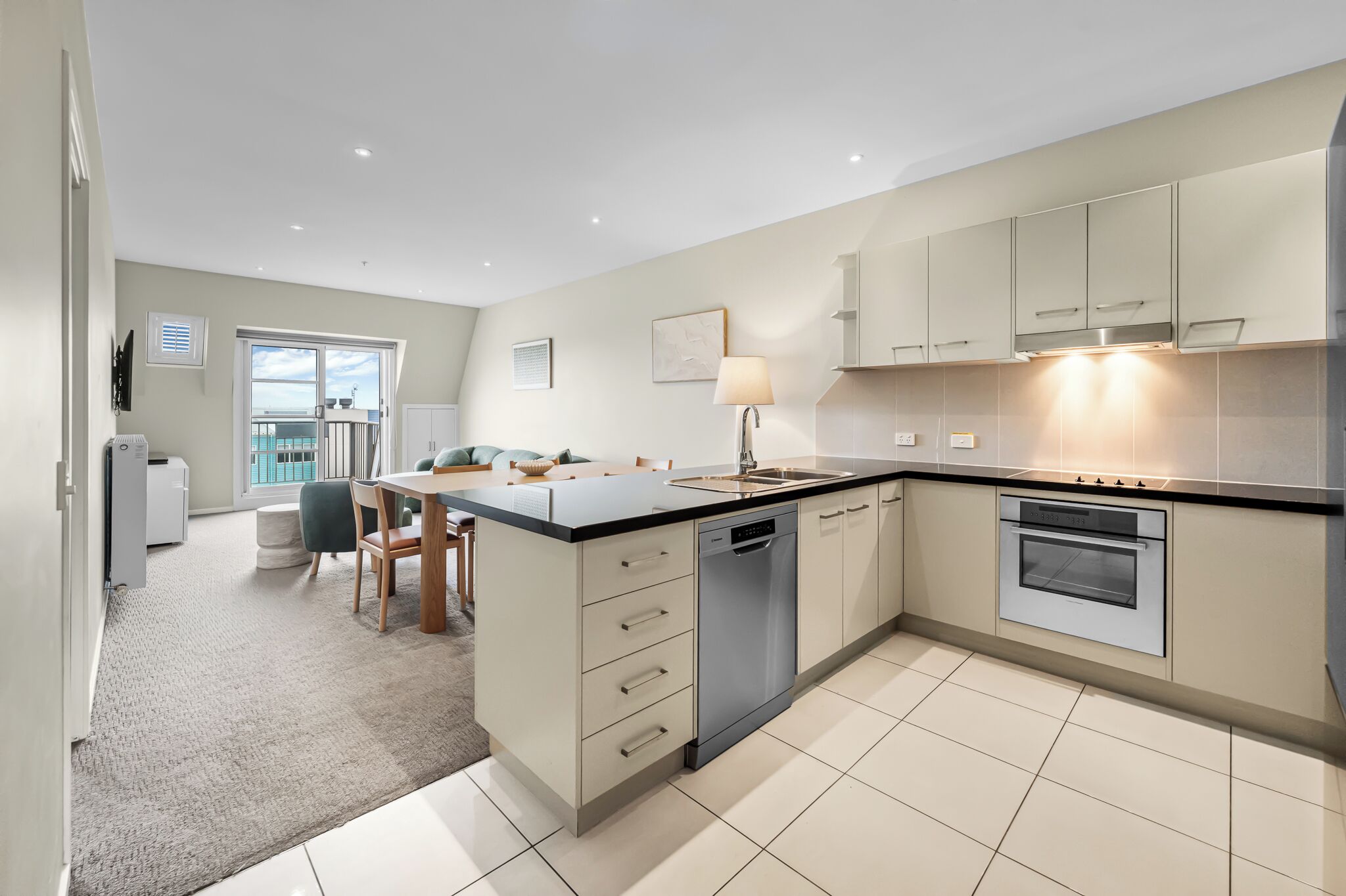 A modern kitchen with a central island, built-in oven, and dishwasher. There is a dining area with a table and chairs, and a view of the outdoors through a large window.