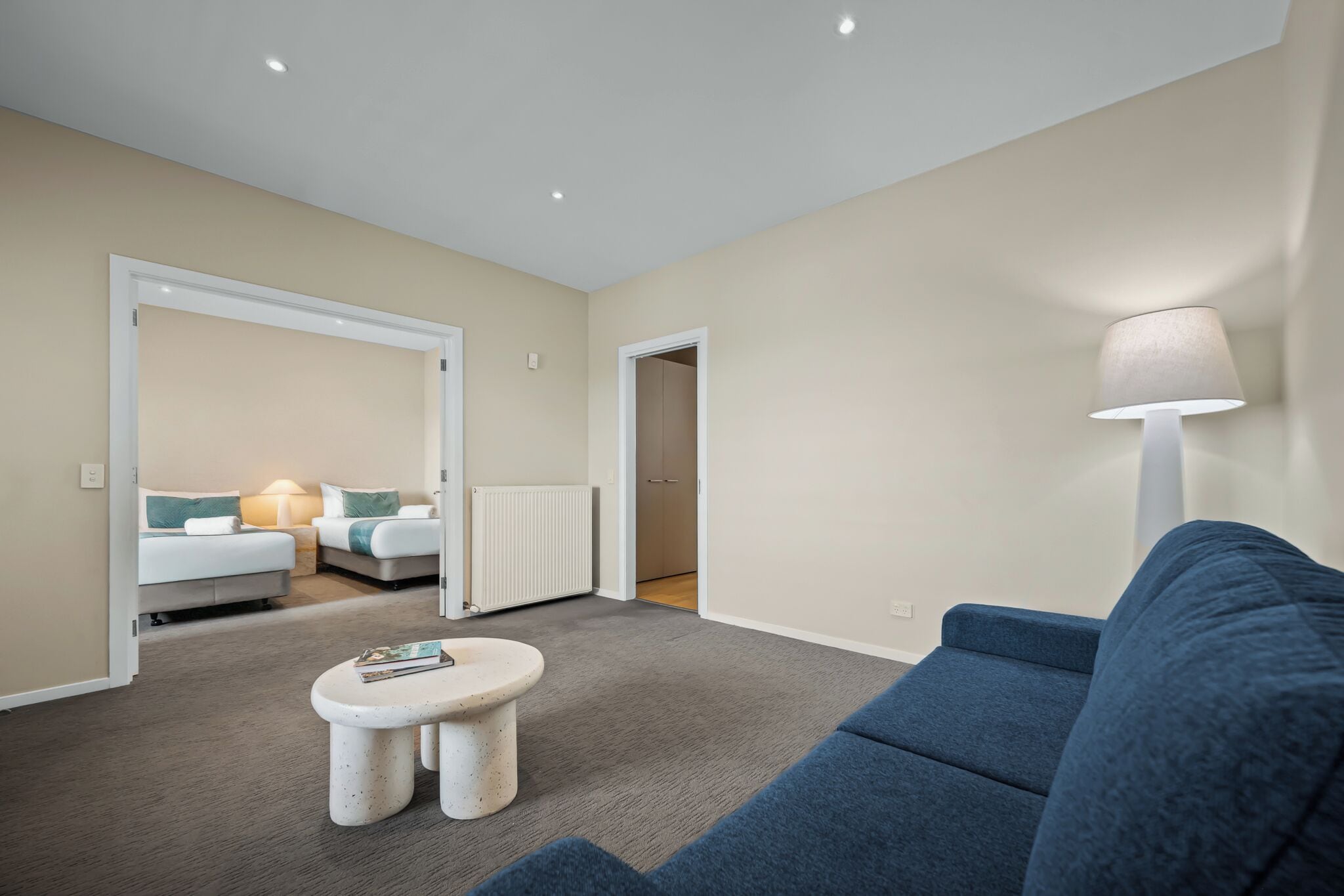 Una habitación de hotel moderna con un sofá azul, una mesita redonda y una lámpara. Se ve una puerta que da a un dormitorio con una cama y un radiador.