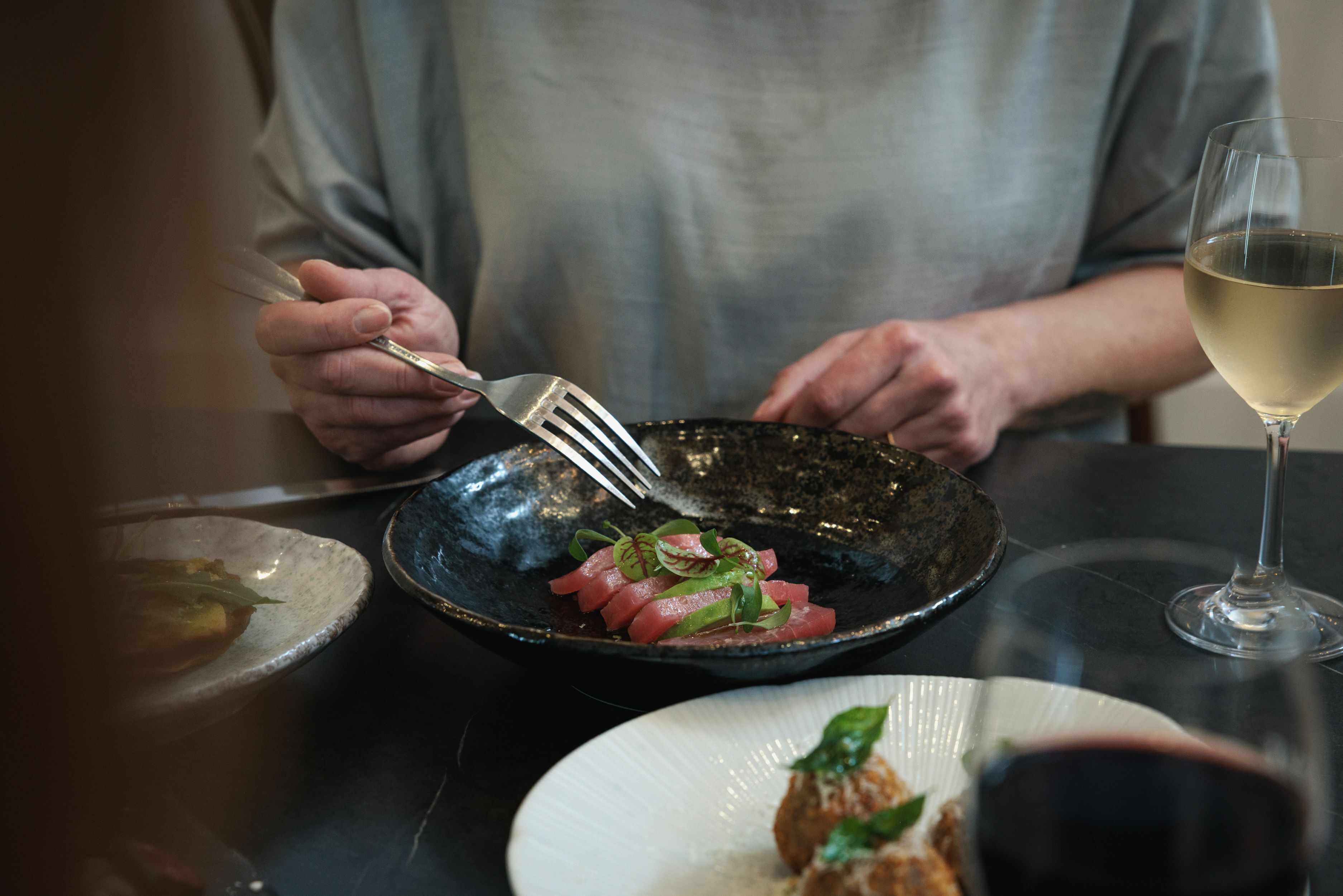 A person dining with a fork and knife, a glass of white wine, and a bowl of food on a table.