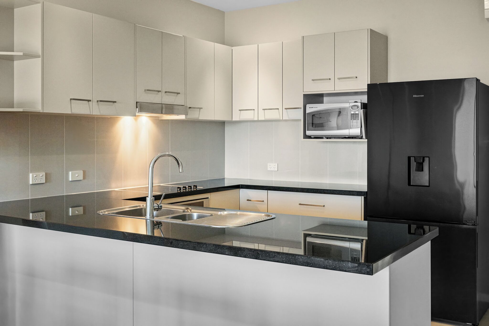 Una cocina moderna con armarios blancos, un lavamanos de acero inoxidable, un refrigerador negro y un microondas integrado.