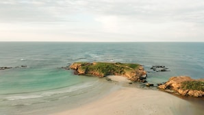Paisagem costeira com uma praia arenosa, afloramentos rochosos e mar calmo.
