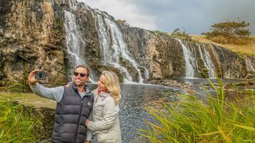 A couple taking a selfie in front of a waterfall with a rocky backdrop and a river in the foreground.