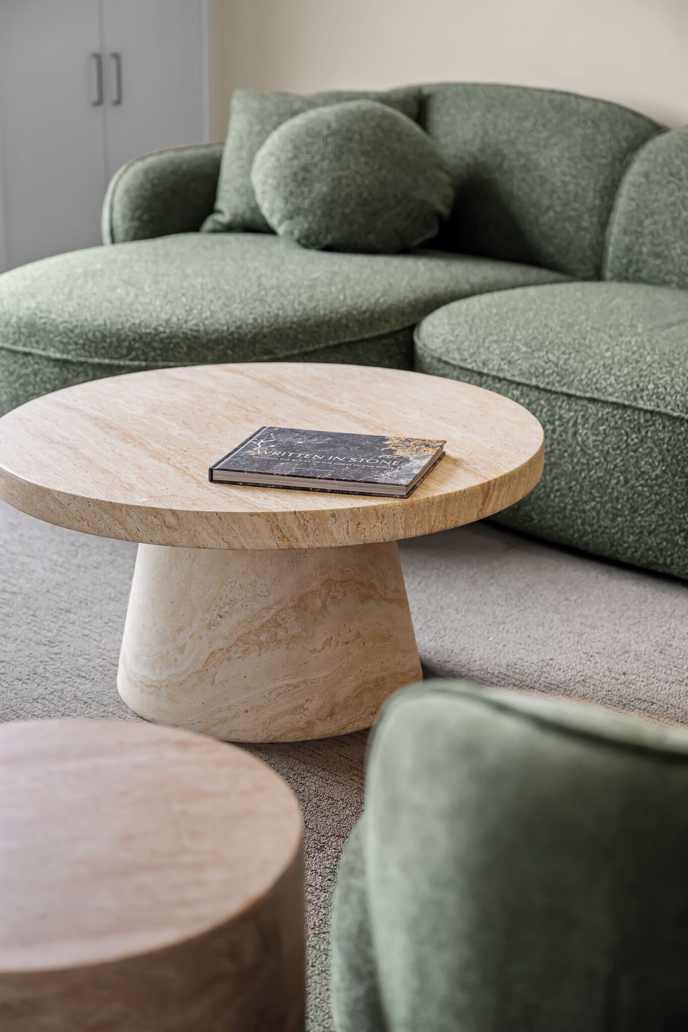 A round marble table with a book on it, placed in front of a green sofa with cushions.