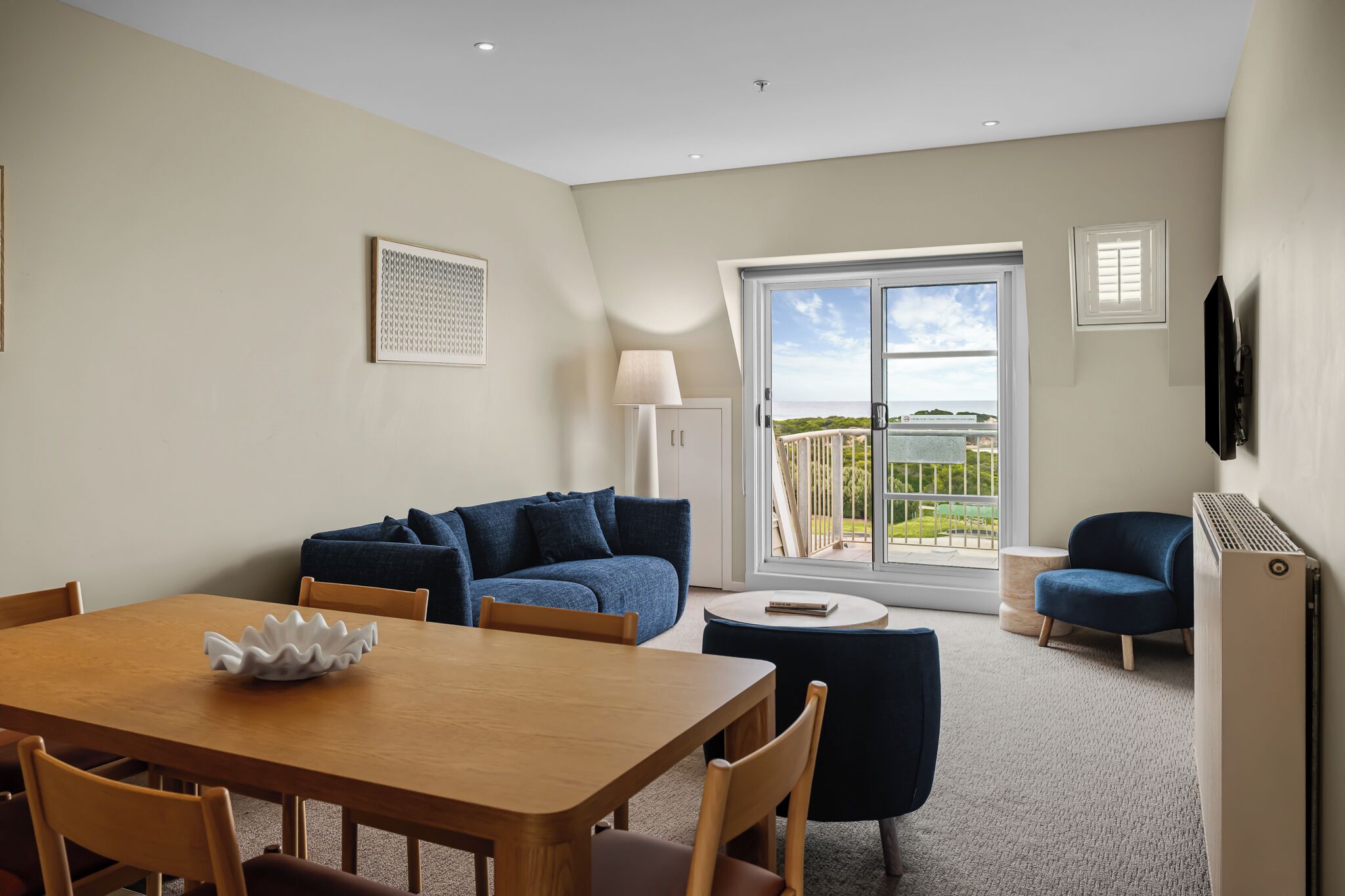 A modern living room with a wooden dining table, blue sofa, and a view of the outdoors through a sliding glass door.