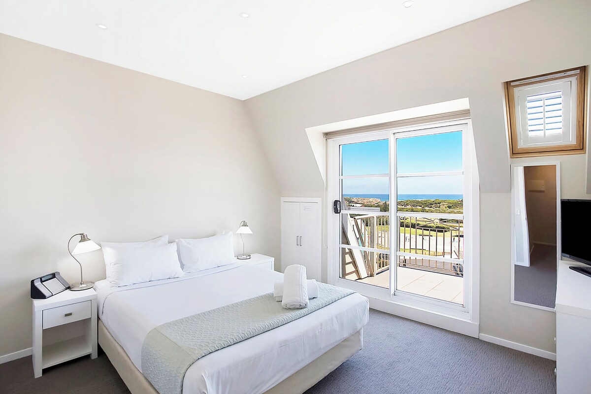 A hotel room with a bed, nightstands, a desk, and a large window with a view.