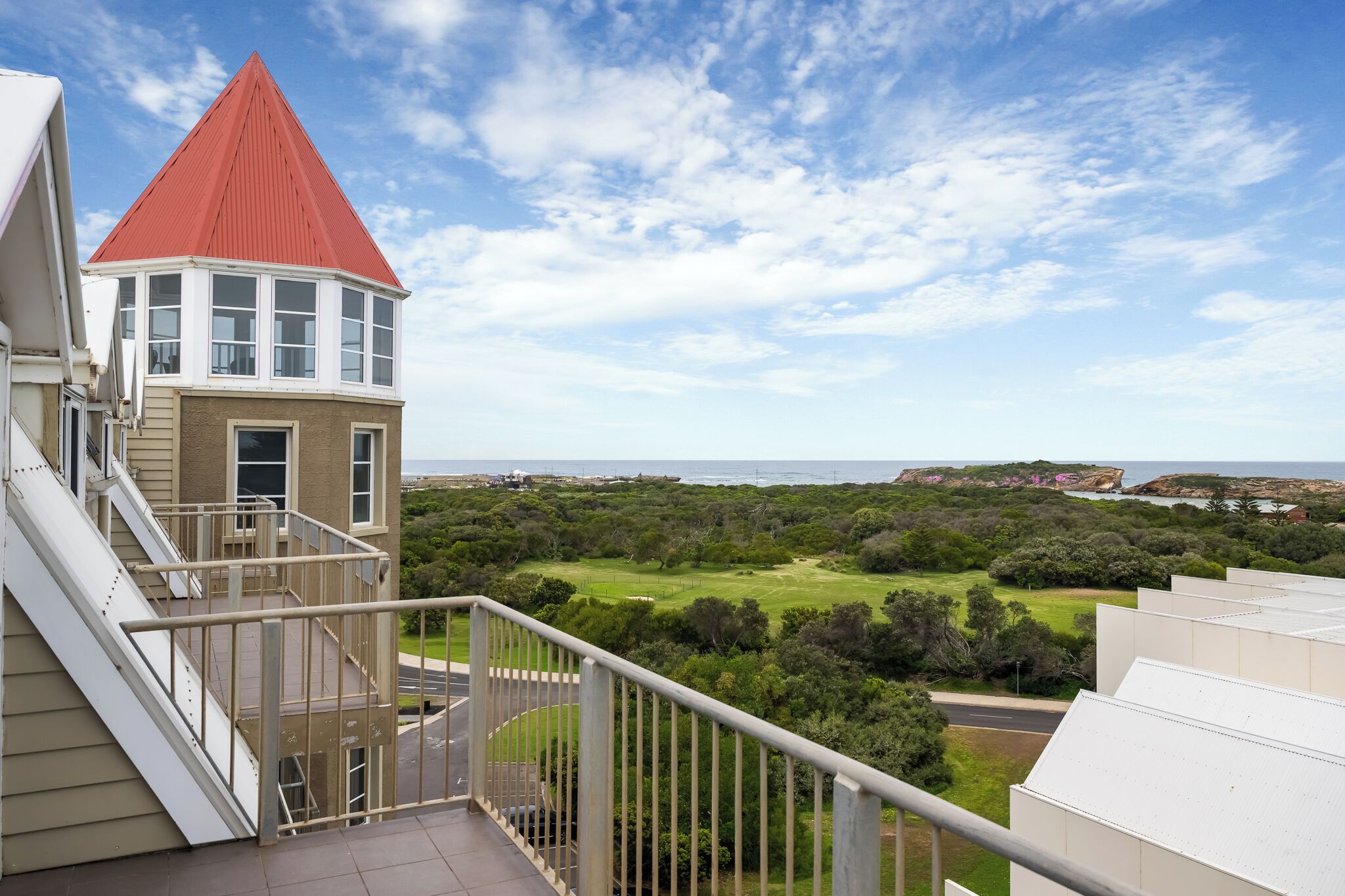 A residential building with a red-roofed tower, a balcony with a railing, and a view of a green landscape and coastline.