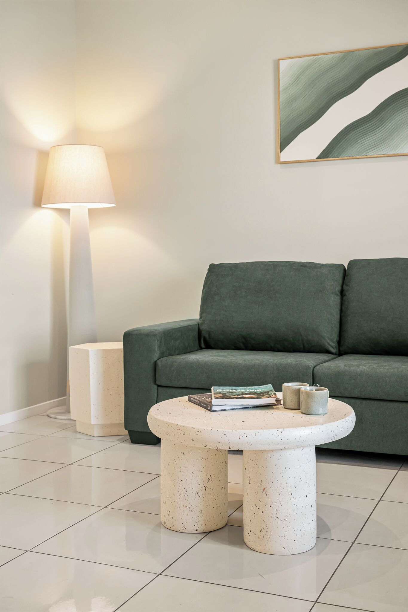Un salon moderne avec un canapé vert, une table ronde ornée de livres et d’une bougie, et un lampadaire.