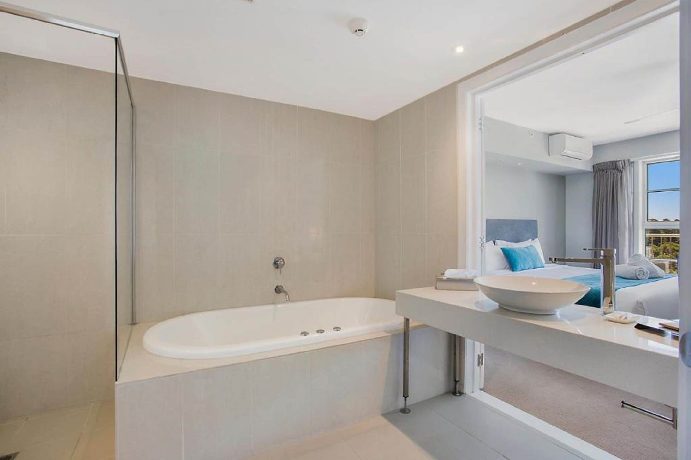 A modern bathroom with a freestanding bathtub, a large mirror, and a visible bedroom with a bed and window.