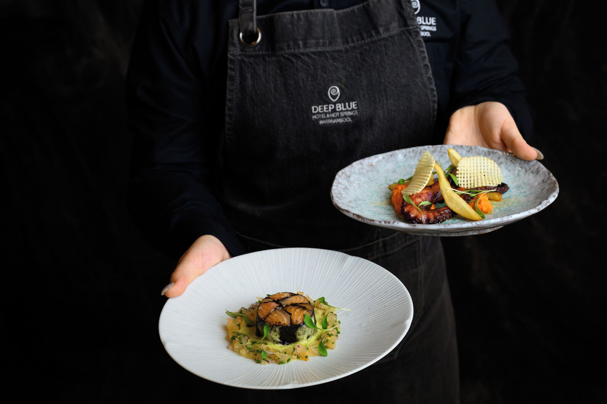 A person holding two plates of food, one with a fish dish and the other with a variety of vegetables and a waffle.