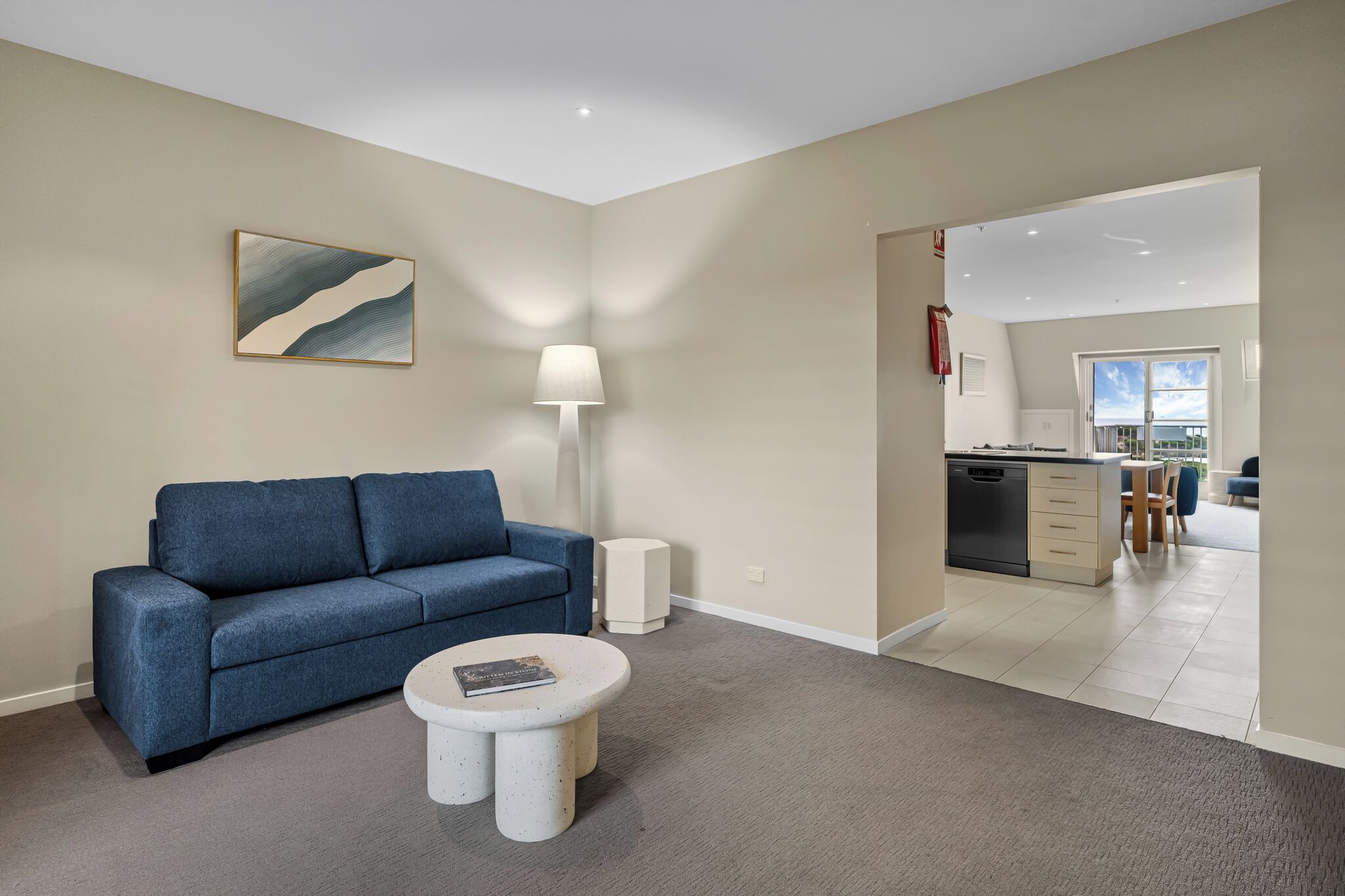 A modern living room with a blue sofa, a round coffee table, and a floor lamp. There is a kitchen visible through an open doorway.