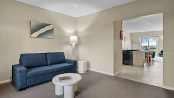 A modern living room with a blue sofa, a round coffee table, and a floor lamp. There is a kitchen visible through an open doorway.