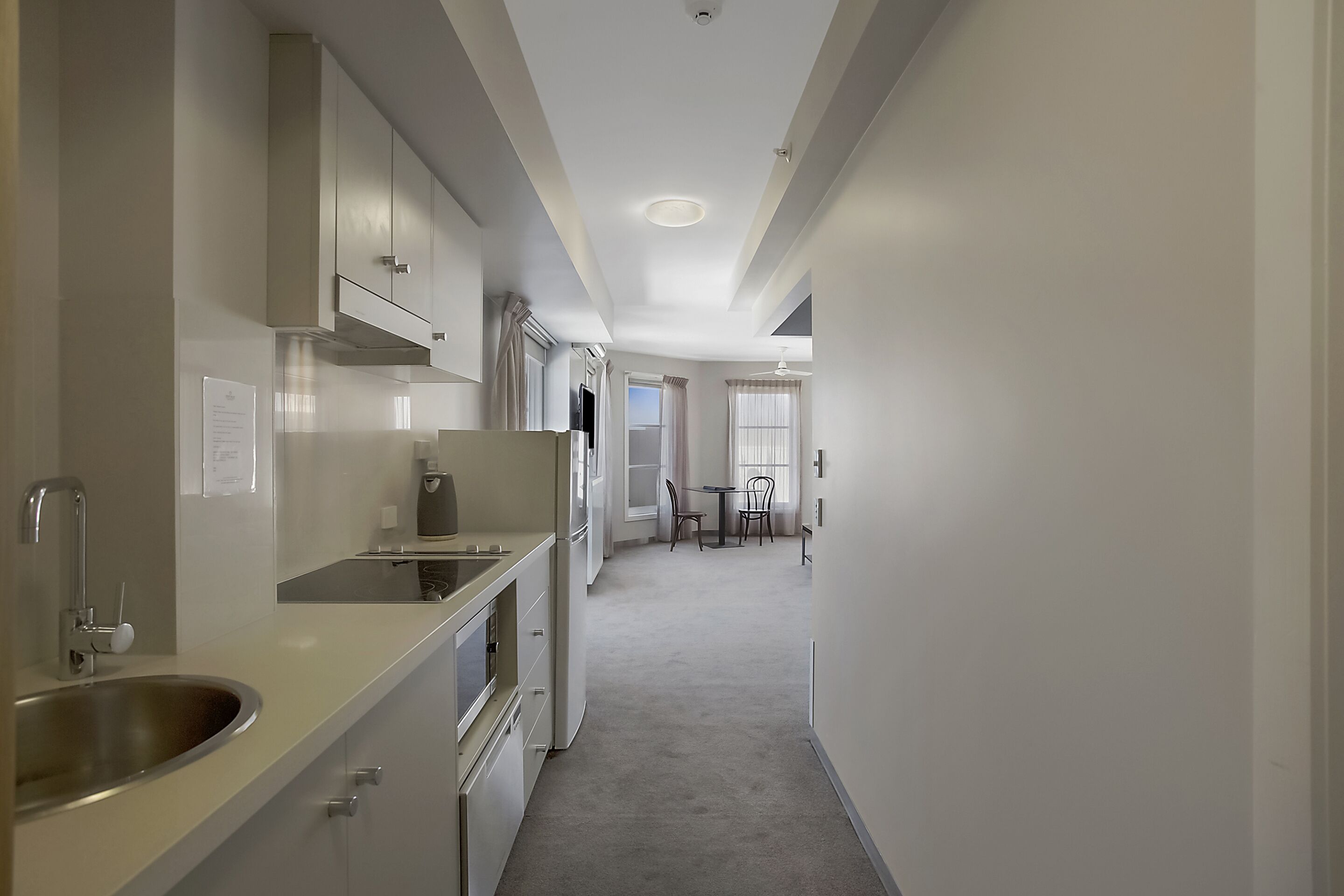 A narrow, well-lit hallway with a kitchen on one side, featuring stainless steel appliances and a sink. A dining area with a table and chairs is visible in the distance.