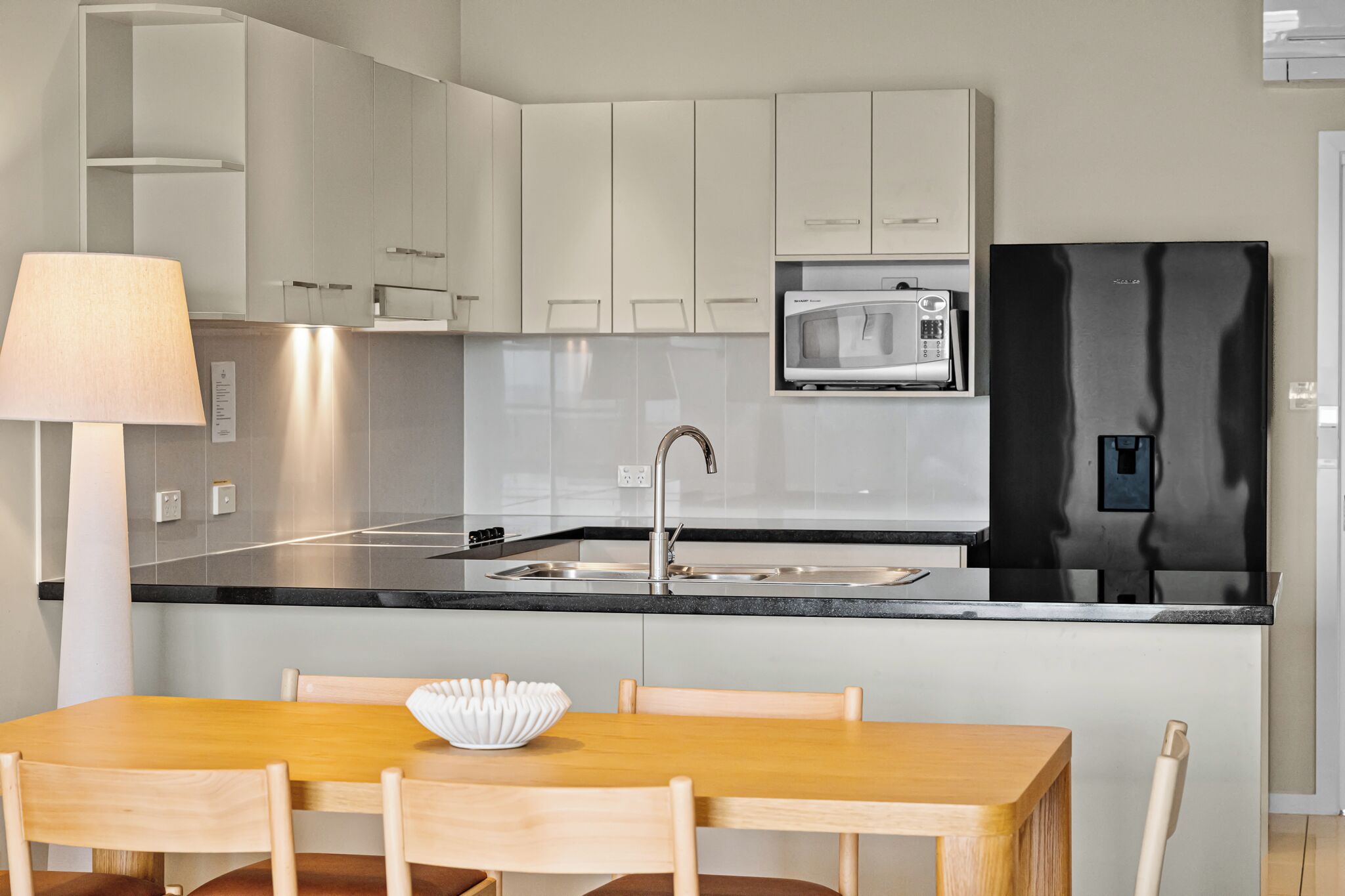 Una cocina moderna con armarios blancos, un refrigerador negro, un microondas y un lavamanos. Hay una mesa de comedor de madera con sillas y un tazón blanco encima.