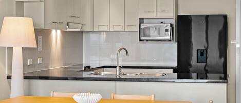 Una cocina moderna con armarios blancos, un refrigerador negro, un microondas y un lavamanos. Hay una mesa de comedor de madera con sillas y un tazón blanco encima.